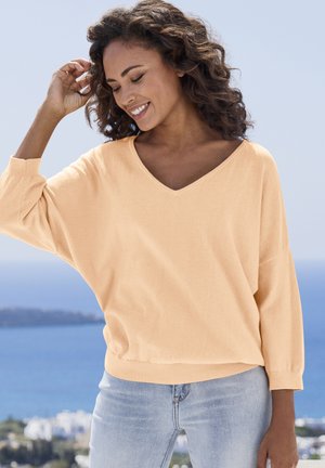 Femme souriante aux cheveux bouclés portant un pull pêche et un jean bleu clair, avec l'océan et une ville côtière en arrière-plan.