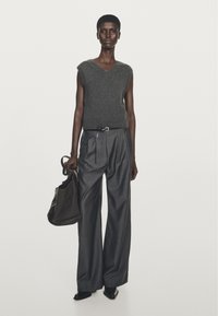 Grey sleeveless knit top with a V-neck, paired with high-waisted, wide-legged dark trousers. Model holds a dark bag, with pointed shoes.