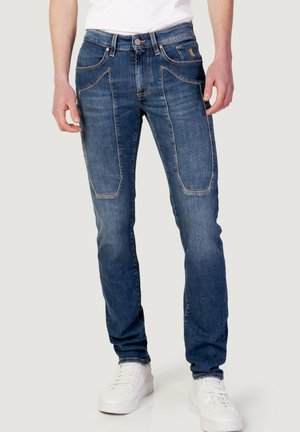 Jeans in denim blu scuro con cuciture a contrasto, tasche frontali con forme uniche e vestibilità slim, abbinati a sneakers bianche.