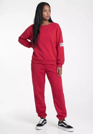 Młoda kobieta z długimi czarnymi włosami, ubrana w czerwoną bluzę i pasujące spodnie dresowe, czarno-białe buty sportowe, stoi z jedną ręką na biodrze.