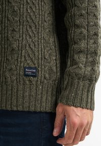 Oliwkowy sweter wykonany na drutach, z teksturowanym wzorem w warkocze i ściągaczami na rękawach. Posiada czarną metkę z logo "Petrol Ind.".