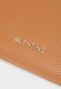 Portafoglio in pelle color tan con superficie texturizzata e logo "VALENTINO" in oro in rilievo. La prospettiva ravvicinata evidenzia i dettagli del materiale.