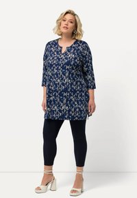 Granatowa tunika z geometrycznym białym wzorem, rękawy do łokcia, zaokrąglony dekolt, w połączeniu z ciemnymi legginsami i białymi sandałami na słupku.