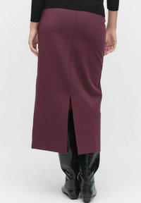 Femme portant une jupe prune mi-longue avec une fente à l'arrière et des bottes noires texturées jusqu'aux genoux, se tenant devant un fond uni.