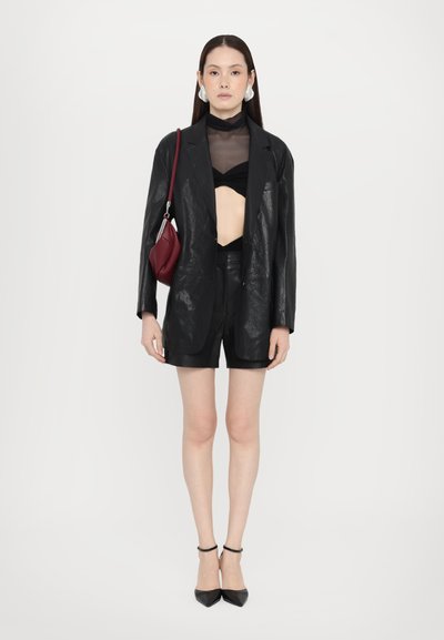 Blazer din piele sintetică neagră cu un finisaj lucios, asociat cu un top negru transparent și pantaloni scurți din piele. Look-ul este completat de tocuri negre ascuțite și o geantă burgundică.