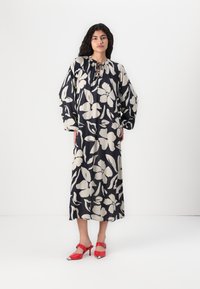 Maximekleid mit navyfarbener Basis und großem cremefarbenem Blumenmuster; verfügt über einen Kordelzug-Ausschnitt und lange, luftige Ärmel. Kombiniert mit roten High Heels.