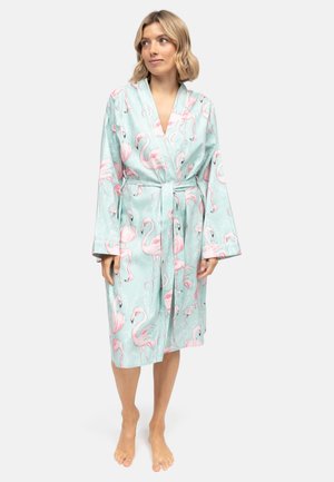 Femme portant une robe légère bleu clair jusqu'aux genoux avec un imprimé flamant rose, nouée à la taille, debout pieds nus sur un fond uni.