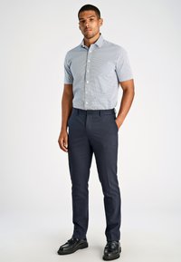Camicia a maniche corte blu con motivo e bottoni bianchi, abbinata a pantaloni eleganti blu navy e scarpe nere, in piedi su uno sfondo neutro.