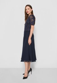 Abito blu navy con top in pizzo e maniche corte, design a trapezio fluido, abbinato a scarpe con tacchi alti nere. Tessuto liscio.