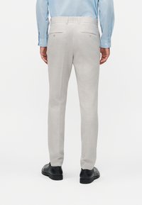 Homme portant un pantalon sur mesure gris clair, une chemise habillée bleu clair et des chaussures habillées noires, debout de dos sur un fond uni.