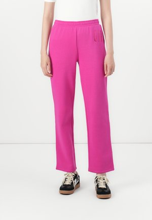 Person iført lyserøde sweatpants med elastisk talje og sorte sneakers med hvide snørebånd, stående mod en hvid baggrund.