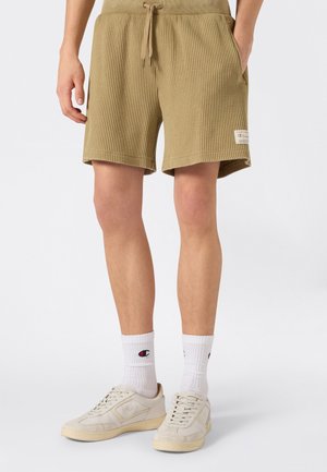 Uomo che indossa pantaloncini beige a coste con coulisse, calzini Champion bianchi e sneaker bianco sporco su sfondo grigio uniforme.