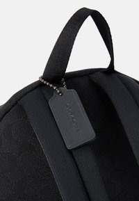 Sac à dos en tissu noir avec un motif texturé, des bretelles ajustables et une petite étiquette attachée par une chaîne à perles.