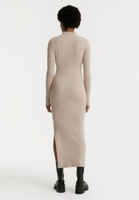 Lang, aangesloten, geribbelde jurk in beige, met een hoge hals en een zijsplits, gecombineerd met zwarte plateau laarzen. Glad textuur, elegante ontwerp.