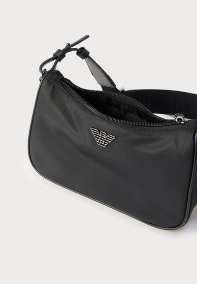 Μαύρη κομψή τσάντα ώμου με ασημί λογότυπο αετού, κλείσιμο με φερμουάρ και ρυθμιζόμενο ιμάντα σε λευκό φόντο.