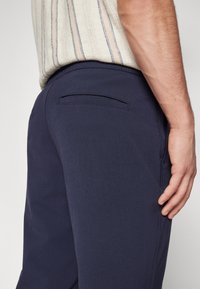 Laivastonsiniset housut, jotka on valmistettu sileästä materiaalista, joissa on yksittäinen takatasku, siistit linjat ja räätälöity istuvuus, esitetty sivulta.