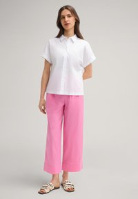 Witte korte mouwen overhemd met knopen, gecombineerd met losse roze broek en crèmekleurige sandalen met zwarte tekst. Soepele texturen, losse pasvorm.