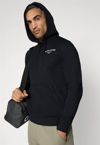 Sweatshirt preto com capuz e bolso frontal, apresentando texto de logótipo branco. O modelo segura uma pequena bolsa cinza com fecho, vestindo calças de tom claro.