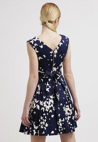 Navy dress with white butterfly print, sleeveless, V-neck back, pleated skirt, and a tie at the waist. Smooth fabric with a fitted bodice.