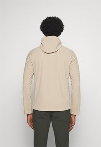 Homme aux cheveux bouclés foncés portant une veste à capuche beige et un pantalon vert foncé, vu de dos sur un fond uni.