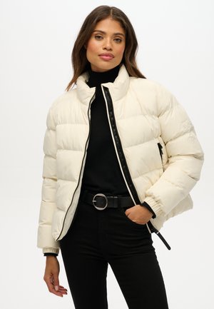 Pufferjack in crème met zwarte ritsdetails, hoge kraag en elastische cuffs. Gestyled met een zwarte coltrui en een zwarte riem.