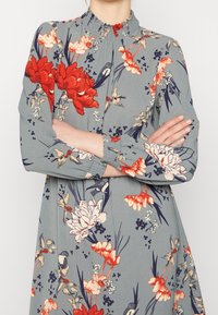 Vestido floral con un cuello alto y fruncido. Presenta un fondo gris, flores rojas y crema, y mangas largas con puños elásticos.