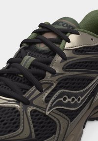 Κοντινό πλάνο μαύρου και ελιάς αθλητικού παπουτσιού με ύφασμα διχτυού, μαύρα κορδόνια και λογότυπο στο πλάι και στη γλώσσα.