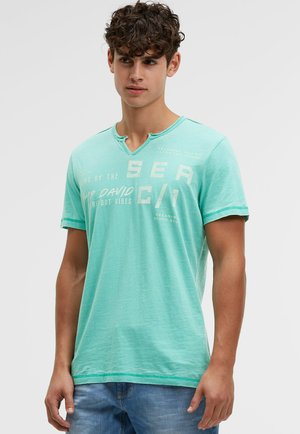 Junger Mann trägt ein helltürkisfarbenes T-Shirt mit Grafik, kurzen Ärmeln und blaue Jeans, steht vor einem einfarbigen grauen Hintergrund.