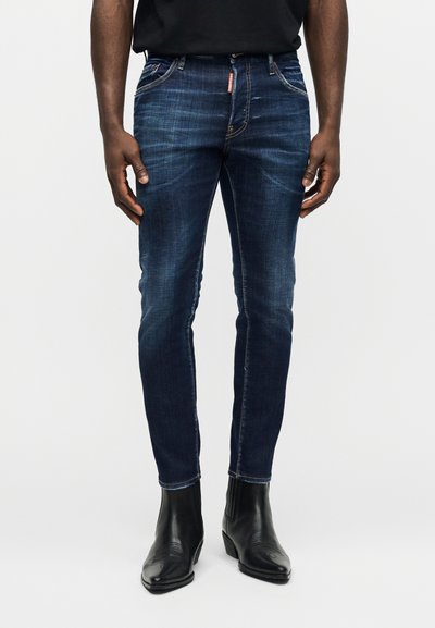 Homme portant un jean slim bleu marine et des bottines en cuir noir, posé devant un fond clair uni.