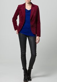 Burgundy kordflorsblazer med enkel knäppning, över en blå topp och svarta byxor i konstläder, kombinerat med svarta snörskor.