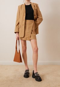 Blazer camel à épaules structurées, haut noir côtelé, mini-jupe camel à ourlet effiloché, et chaussures plateformes noires. Comprend un sac à main marron.