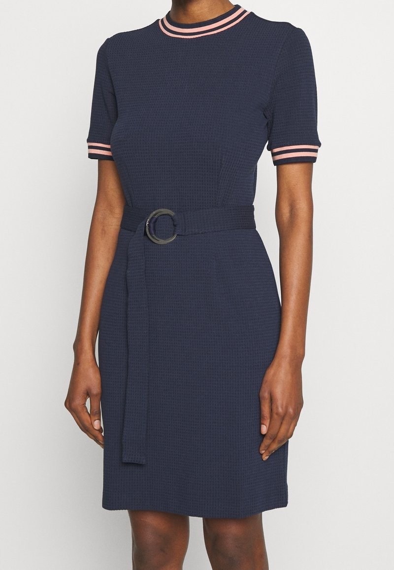 Robe bleu marine à manches courtes avec un bord rayé rose sur les manches et le col, taille ceinturée avec une boucle ronde, portée par une personne debout.