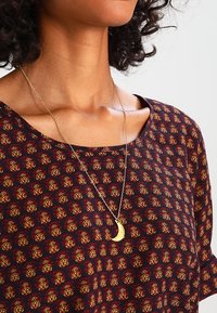 Kvinna som bär en mönstrad topp i röd och gul färg samt ett guld crescentmånehänge på ett tunt kedjehalsband.