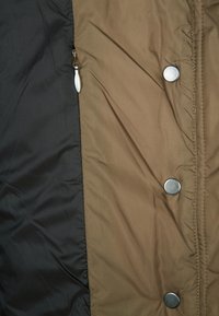 Černá a hnědá látka bundy se stříbrnými patentními knoflíky a malým zipem na černé straně.