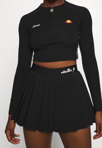 Svart långärmad crop top med dragkedjekrage och broderad logotyp. Kombinerad med en plisserad svart kjol med märkesmidja.