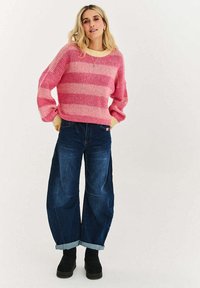 Růžový pruhovaný pletený svetr se žlutými žebrovanými manžetami, kombinovaný s širokými tmavými džínami s mírně vyhrnutými konci a černými kotníkovými botami.