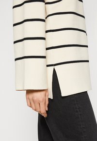 Suéter de rayas en crema y negro, con tejido texturizado, mangas largas y una abertura lateral cerca del dobladillo. Combinado con jeans negros.