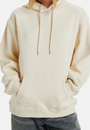 Person iført en off-white hættetrøje med snore og en forlommer, hænder delvist inde i lommen, kombineret med mørke jeans.