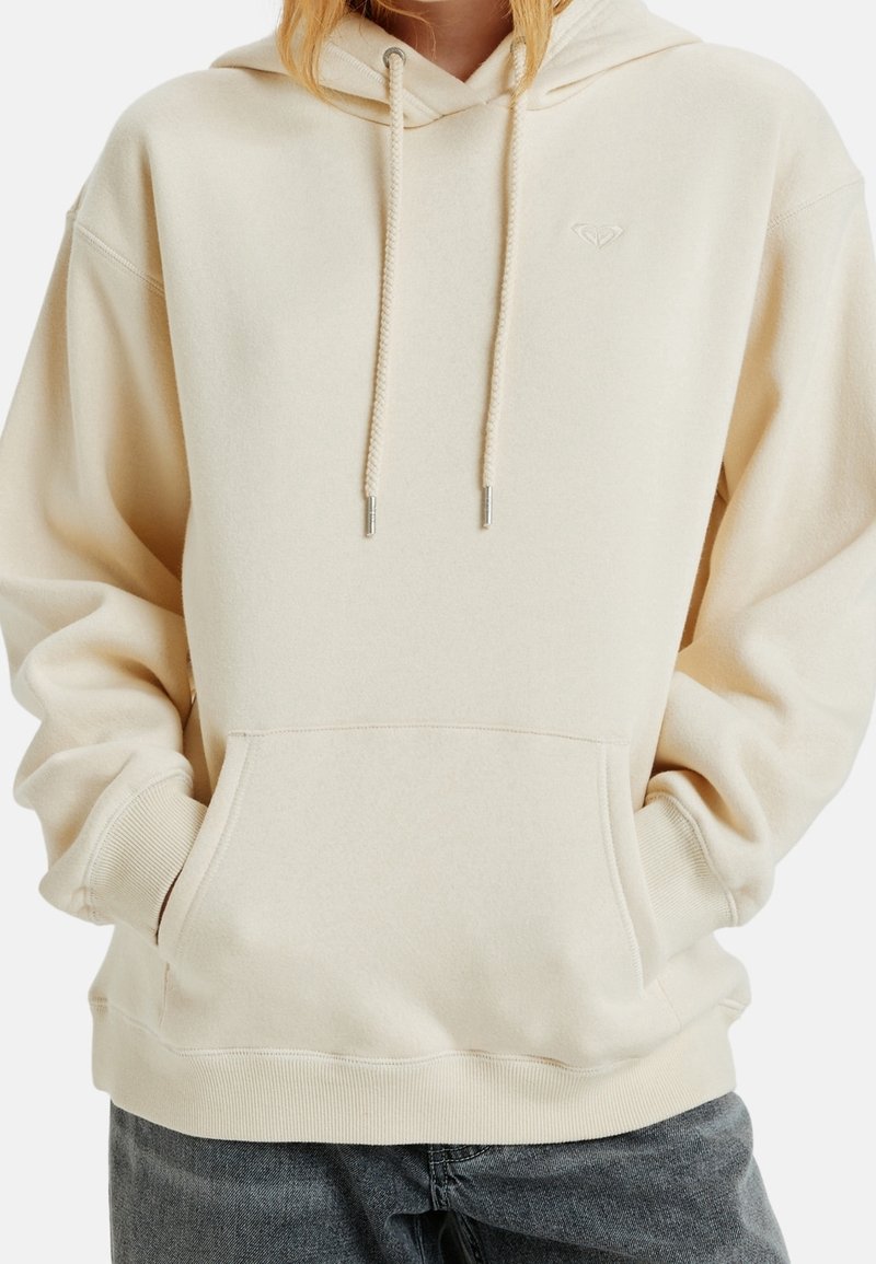 Personne portant un sweat à capuche blanc cassé avec des cordons et une poche avant, les mains partiellement dans la poche, associé à un jean foncé.