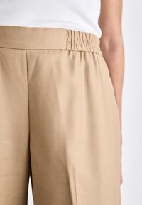 Μπεζ παντελόνι ψηλής μέσης με ελαστική γα gathered, μ�ε λεία υφή και πλαϊνές τσέπες, απεικονίζεται σε ουδέτερο φόντο.