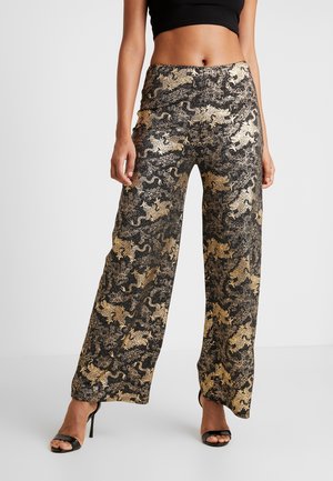 Frau trägt schwarze und goldene, breitbeinige Hosen mit Drachendruck, ein schwarzes, kurz geschnittenes Oberteil und schwarze, hochhackige Sandalen.