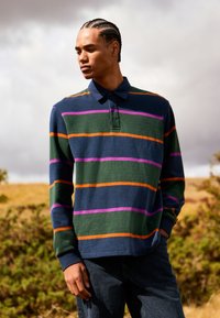 Pruhované polo tričko v námořnické modři, zelené a oranžové barvě s fialovými pruhy. Má límec a dlouhé rukávy, vyrobené z texturované látky.