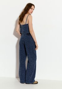 Námořnická modrá pletená overal s širokými nohavicemi, otevřeným designem zad a nastavitelnými popruhy. Disponuje texturovanou tkaninou a vypasovaným pasem.