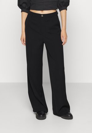 Kobieta ubrana w czarne spodnie z szerokimi nogawkami, czarny krótki top z bufkami na rękawach oraz czarne masywne buty, stojąca na tle jednolitego jasnego tła.