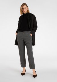 Kobieta w czarnym kożuchu typu shearling, czarnym golfie, szarych spodniach w stylu cargo oraz czarnych szpilkach z zaostrzonym noskiem, stojąca z rękami w kieszeniach.