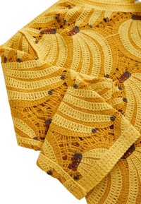 Hæklet gul stof med gentagne bananmotiver i lyse og mørke nuancer, med brune stilke og tekstureret hulmønster i baggrunden.