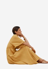 Gelbes, strukturiertes Maxikleid mit kurzen, weiten Ärmeln, tailliertem Schnitt und fließendem Rock. Kombiniert mit flachen Sandalen und schlichten Armbändern.