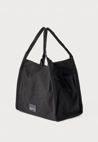 Čierna taška na rameno vyrobená z odolnej tkaniny s dvoma dlhými rúčkami, ozdobným zaviazaným detailom a predným vreckom s nášivkou s logom.
