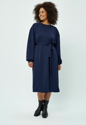 Marineblaue, langärmlige Kleid mit einem taillierten Gürtel. Weicher Stoff, knielang, lässige Passform, mit seitlichen Nahtdetails versehen. Getragen mit schwarzen Knöchelstiefeln.