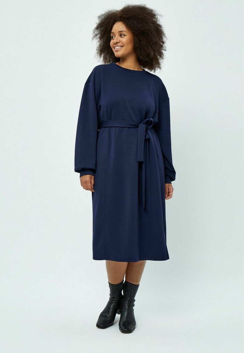 Vestido azul marino de manga larga con cintura ceñida. Tela suave, hasta las rodillas, corte holgado, terminado con detalles en las costuras laterales. Llevado con botas negras de tobillo.